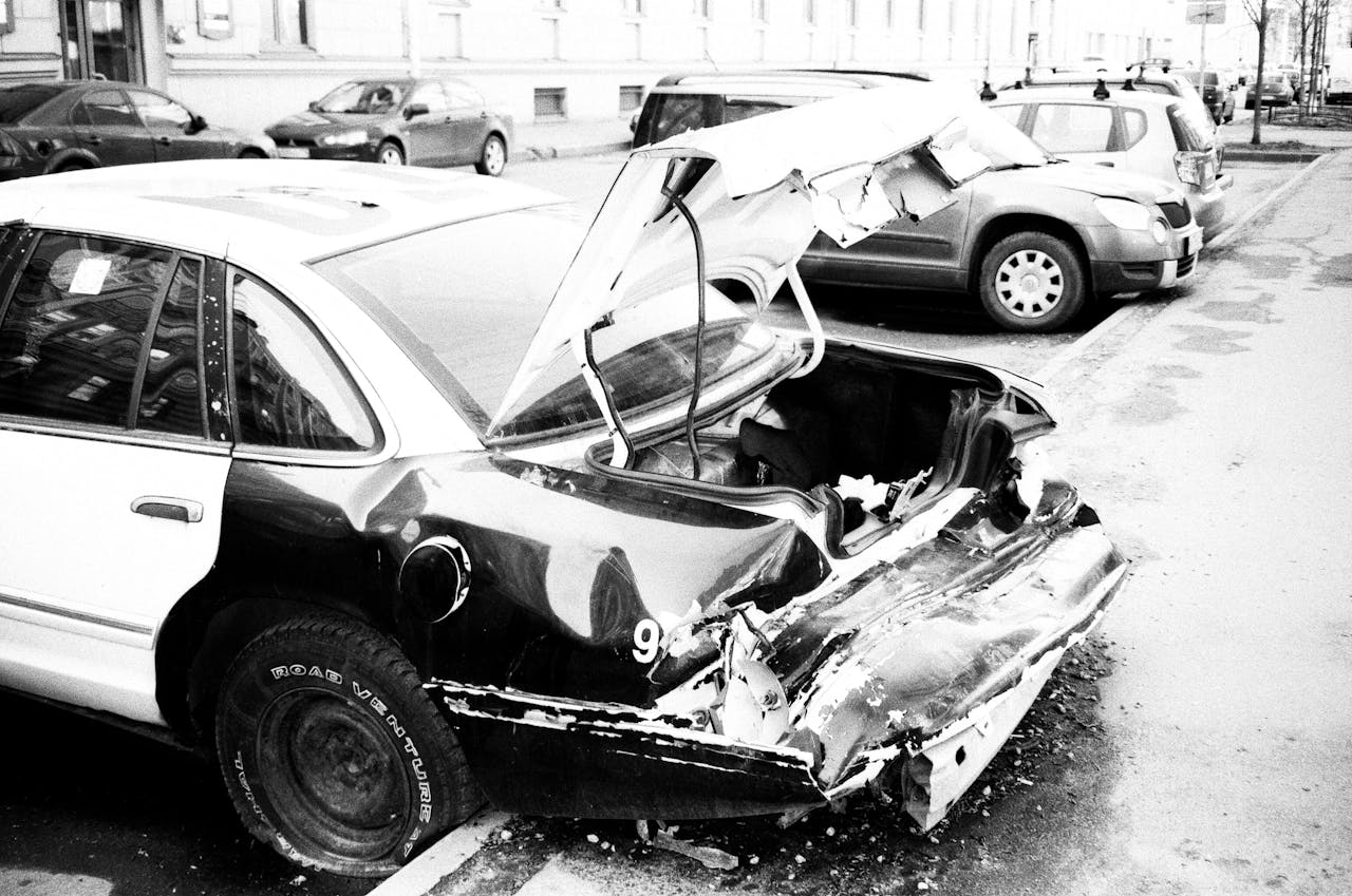 A black and white photo of a wrecked car on an urban street, highlighting vehicle damage.